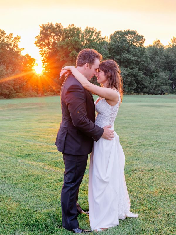 A couple embraces at sunset in a grassy field, dressed in wedding attire.