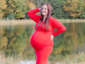 Pregnant woman in a red dress posing by a lake with autumn trees.