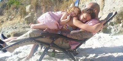 A man and two girls cuddle on a beach chair in a sunny sandy beach setting.