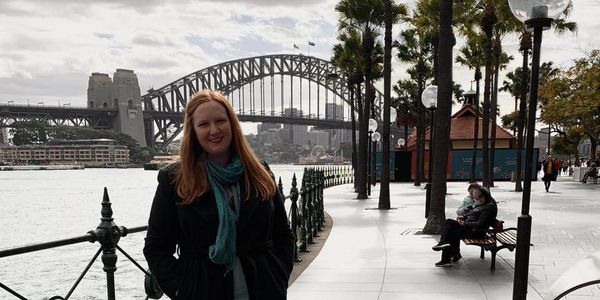 Woman in a black coat standing near Sydney Harbour Bridge on a cloudy day.