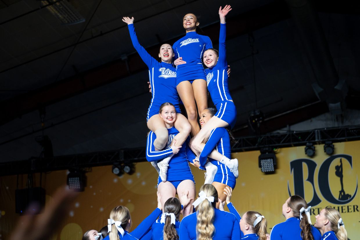 Cheerleaders in blue uniforms performing a high pyramid stunt indoors.