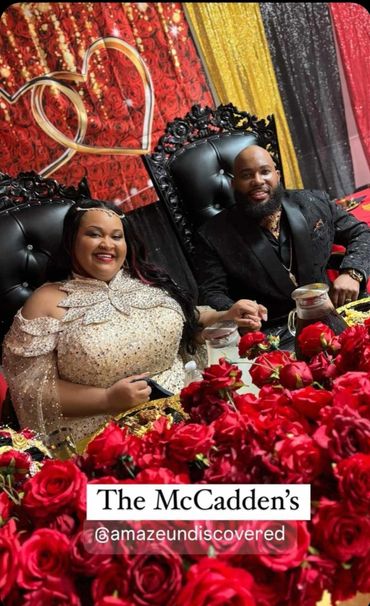 A couple sits at a decorated table with red roses and heart backdrop.