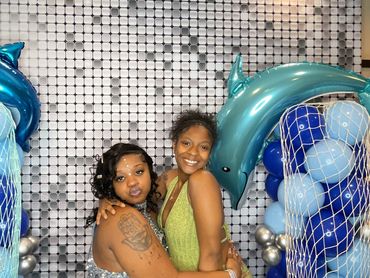 Two women smiling and posing in front of a silver sequin backdrop with blue dolphin balloon arches.