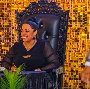 Smiling woman in elegant black dress sitting on a regal chair with golden backdrop.