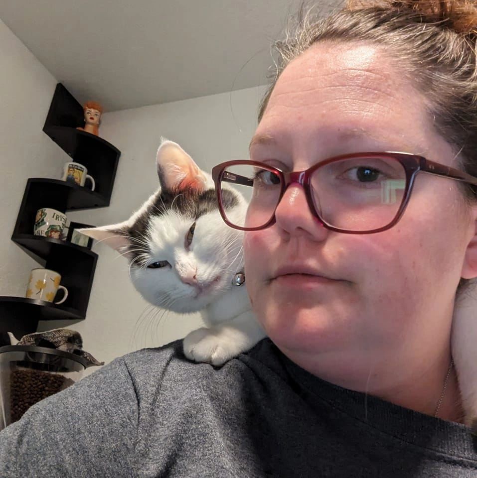 A woman with glasses and a cat resting on her shoulder indoors.