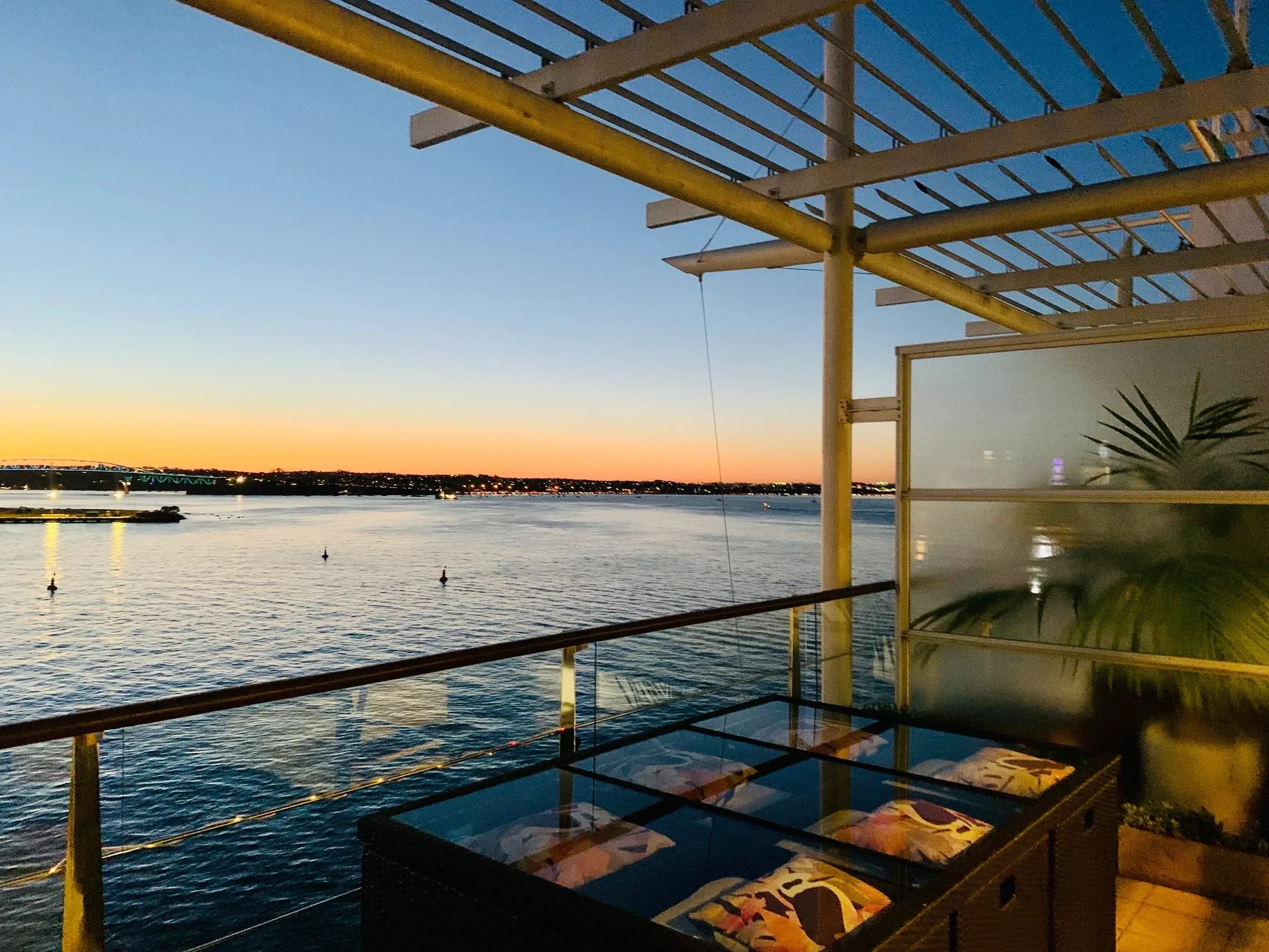 Sunset view over calm waters from a cozy outdoor patio with a glass table and cushions.