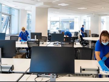 Workers in blue uniforms cleaning desks in a modern office space.