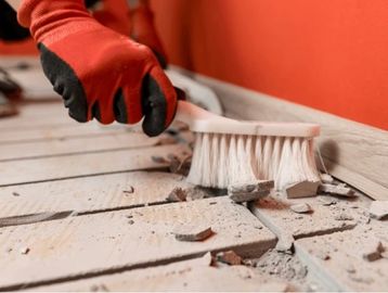 Hand in glove sweeping debris from tiled floor near orange wall.