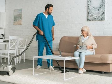 Caregiver vacuuming carpet while elderly woman sits on sofa.