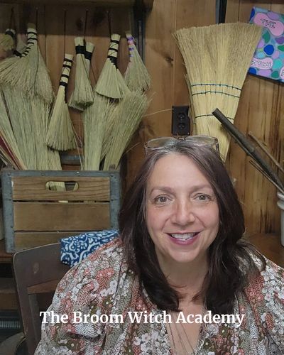 broom making teacher smiling in front of a wall of brooms she has made.