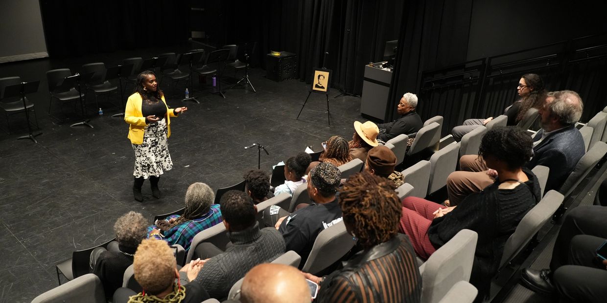 A woman in a yellow jacket speaks to an audience in a theater setting.