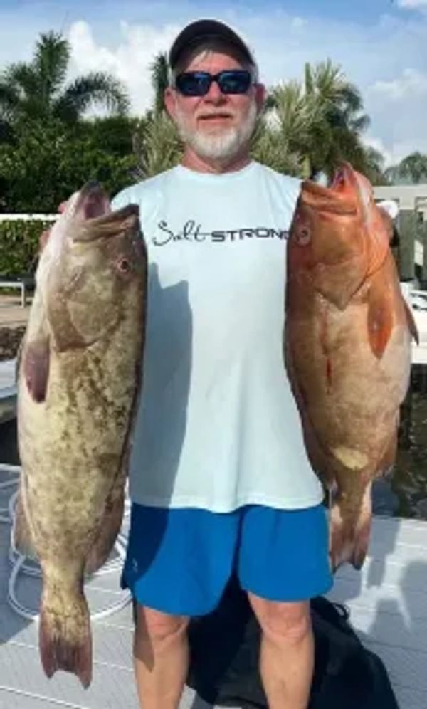 Man holding two large fish, standing outdoors near palm trees.