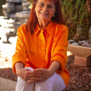 Smiling woman in an orange blouse and white pants sitting outdoors.