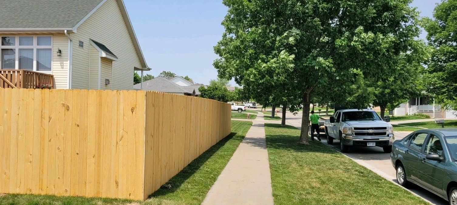 A suburban sidewalk with a wooden fence, trees, and parked vehicles under a clear sky.