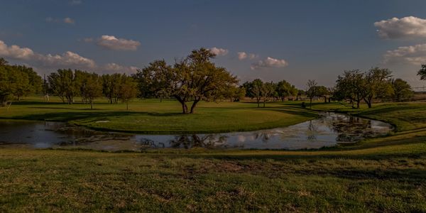 A serene golf course with a water hazard and scattered trees under a blue sky.