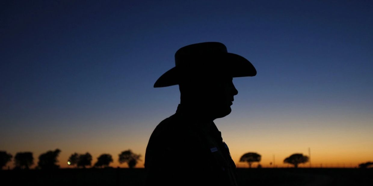Silhouette of a person wearing a cowboy hat at sunset with trees in the background.