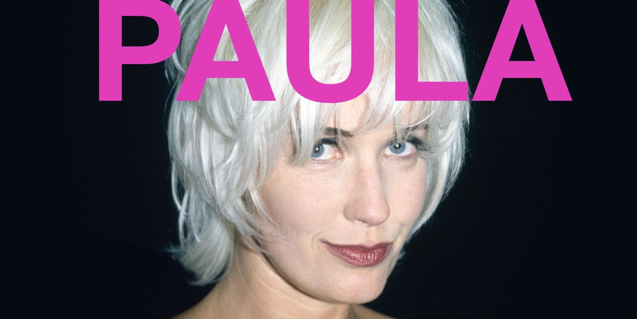 Close-up of a woman with short white hair and pink text reading 'PAULA'