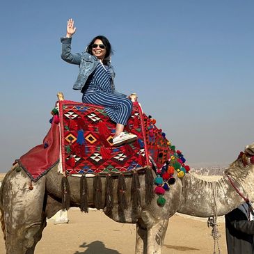 A woman waves happily while sitting on a decorated camel in a desert.