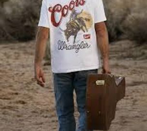 Person in jeans and a Coors Wrangler shirt holding a brown guitar case.