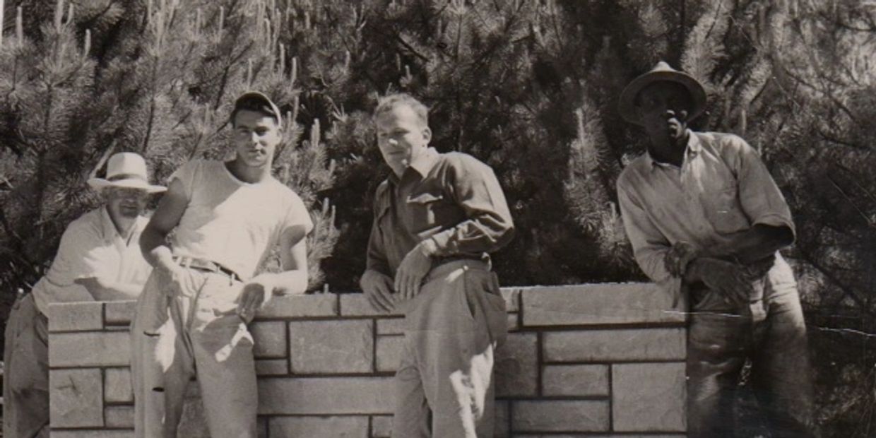 Four men posing by a stone wall under pine trees in a vintage black-and-white photo.