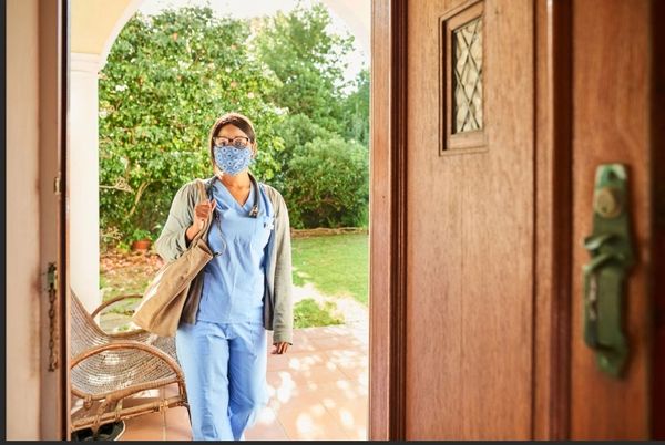 Nurse in scrubs and mask entering a home with a bag and stethoscope.