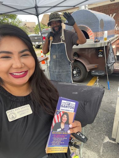 Woman holding campaign flyers with a smiling man waving behind a pizza oven.