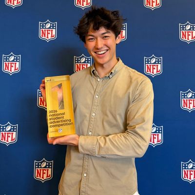 Young man holding 2026 National Student Advertising Competition award with NFL backdrop.