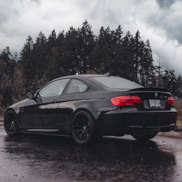Black BMW parked on wet road with forest backdrop under cloudy sky.