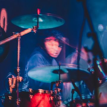 Blurred image of a drummer playing a red drum set under colorful lighting.