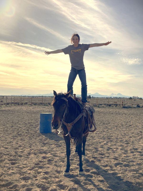 A person balances standing on a horse in an outdoor arena during sunset.