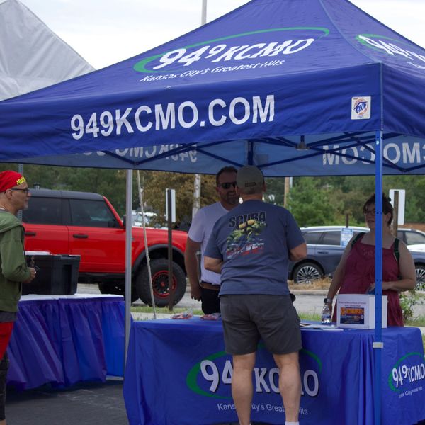 People gather under a 94.9 KCMO promotional tent at an outdoor event.