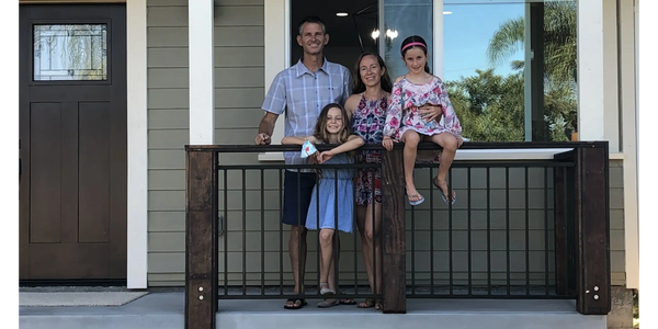 A family of four posing on their front porch with a green house background.