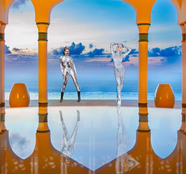 Two models in futuristic outfits pose by a reflective pool with ocean and sky backdrop.