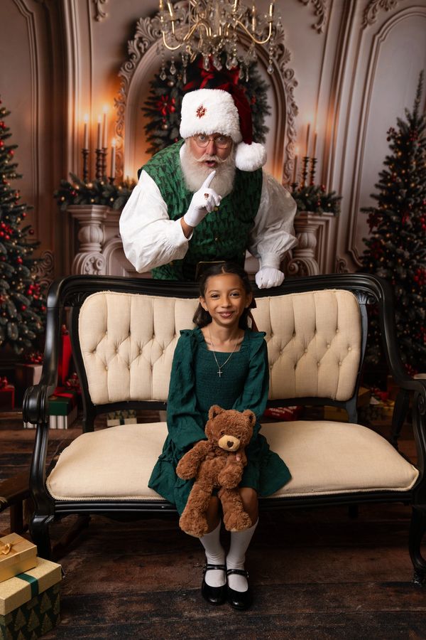 Santa making a shushing gesture behind a smiling girl holding a teddy bear.