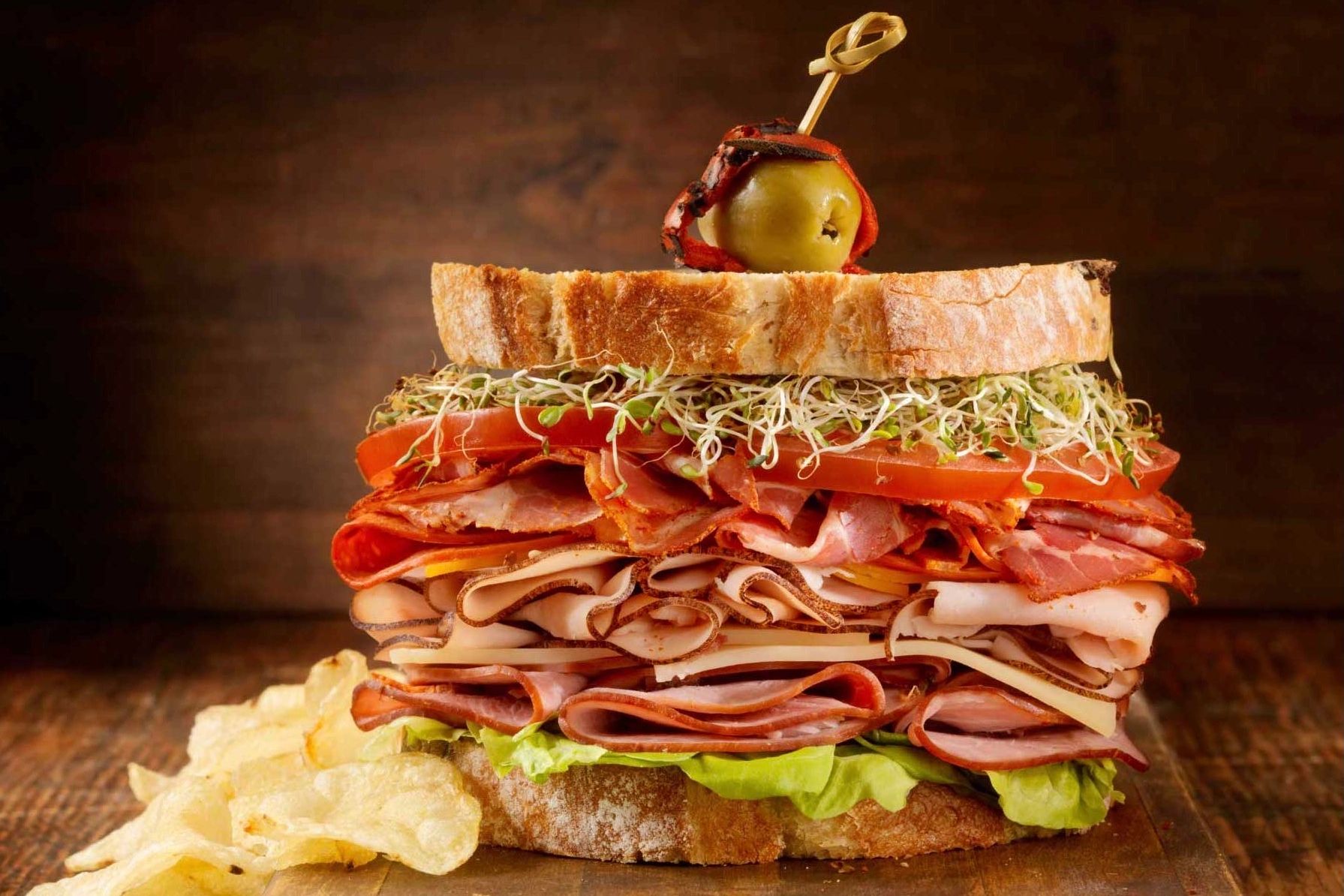 A delicious deli meat and salad sandwich, with crisps and olives on the side.