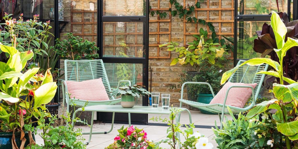 Two garden chairs and a small table on a sunny patio beside brickwork and black-framed glazing.