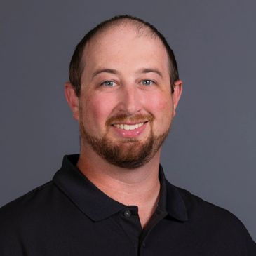 Smiling man with a beard wearing a black polo shirt against a gray background.