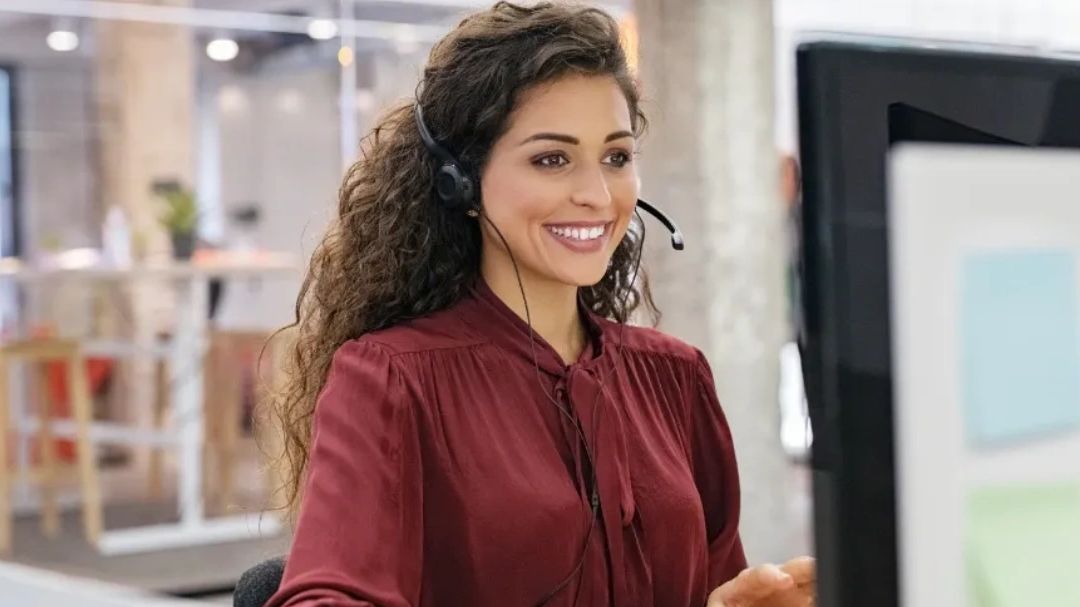 A smiling woman with a headset working at a computer in an office.