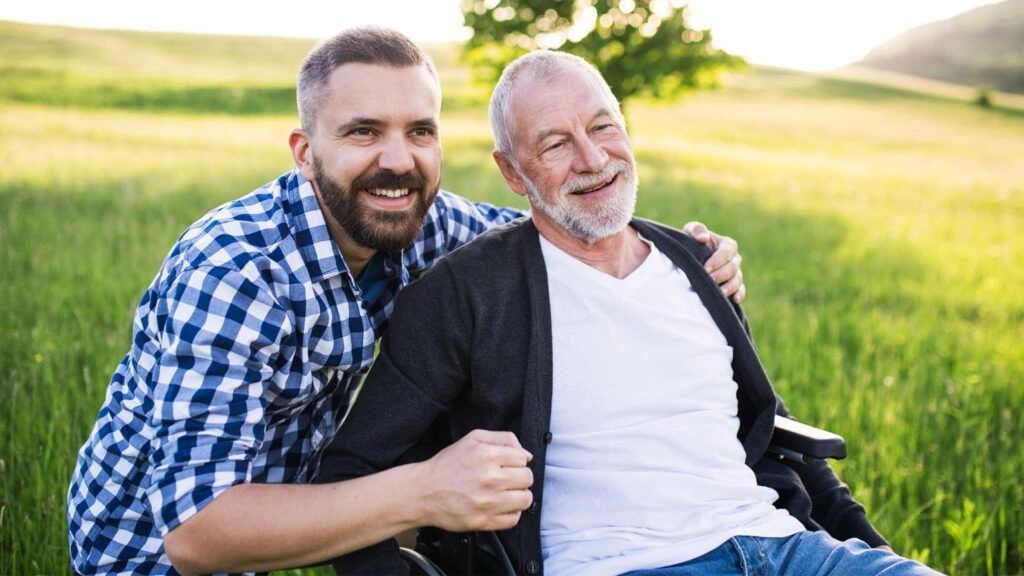 A younger man happily embraces an older man in a wheelchair outdoors.
