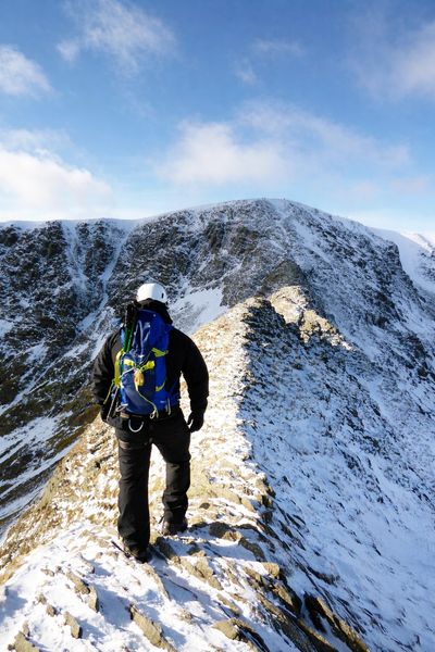 A climber walks along a snowy mountain ridge on a clear day.