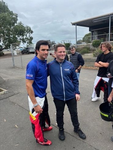 Two men posing for a photo outdoors near a race track.