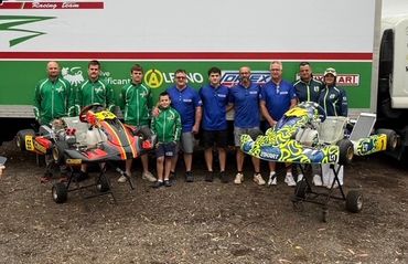 A racing team poses with two colorful go-karts in front of their trailer.
