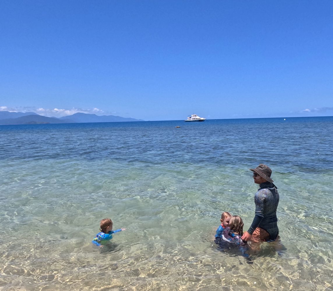 Family enjoys shallow, clear waters under a bright blue sky at the beach.