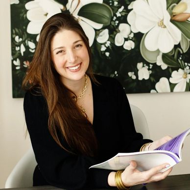 Smiling woman with long hair holding a book, sitting in a chair.