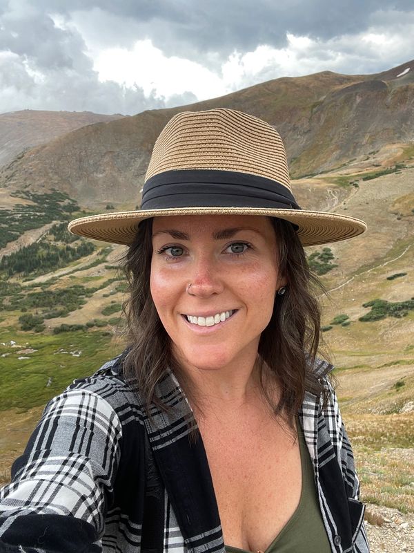 A woman in a hat smiling with mountains in the background.