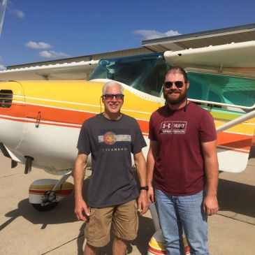 Two men standing in front of a small airplane on a sunny day.