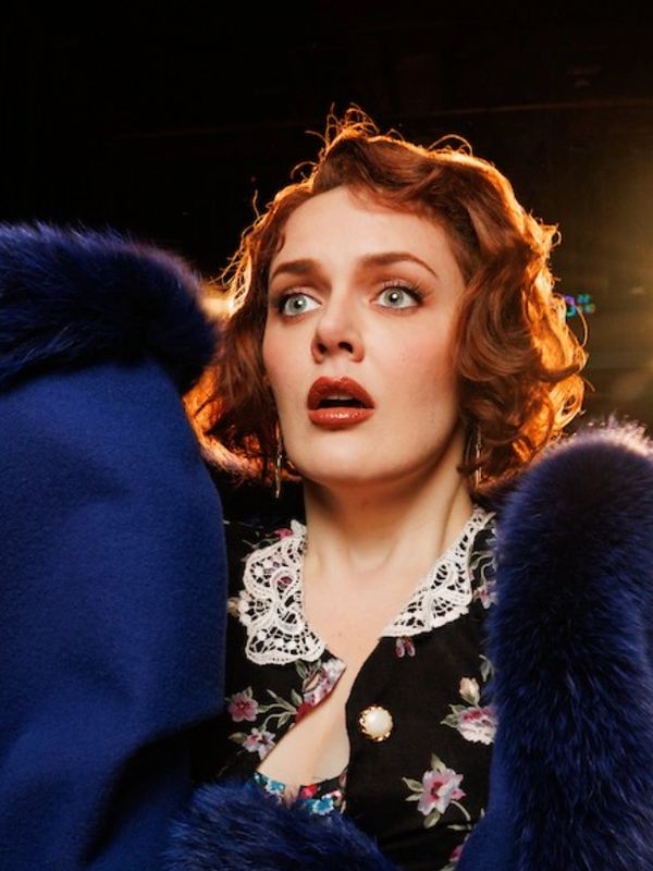 A woman with red hair and a blue fur coat looks surprised on a dimly lit stage.
