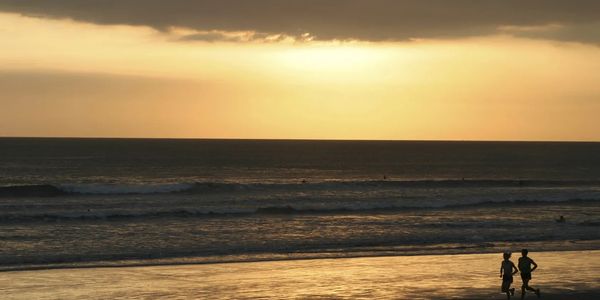 Two people walking along a beach at sunset with surfers in the water.