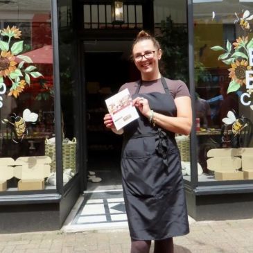 Smiling woman in apron holding a booklet outside a cafe.