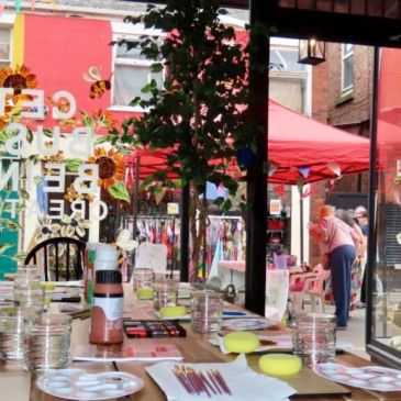 Art supplies on a table inside a cafe with a street market visible outside.